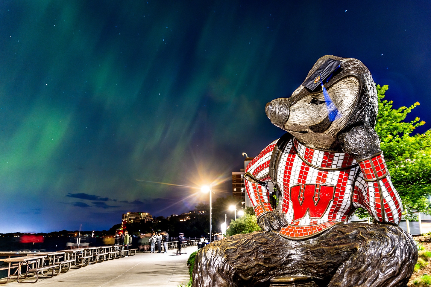 "Well Red," a sculpture by artist Douwe Blumberg of a studious-looking UW-Madison mascot Bucky Badger, is decorated with a Wisconsin School of Business graduation cap as he looks out on the Aurora borealis – commonly called the Northern Lights – as they grace the night sky above Lake Mendota along the University of Wisconsin–Madison shoreline on May 10, 2024, the night before commencement. (Photo by Taylor Wolfram / UW–Madison)