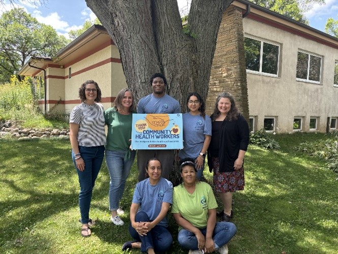 Seven person group standing in front of tree on a sunny day.