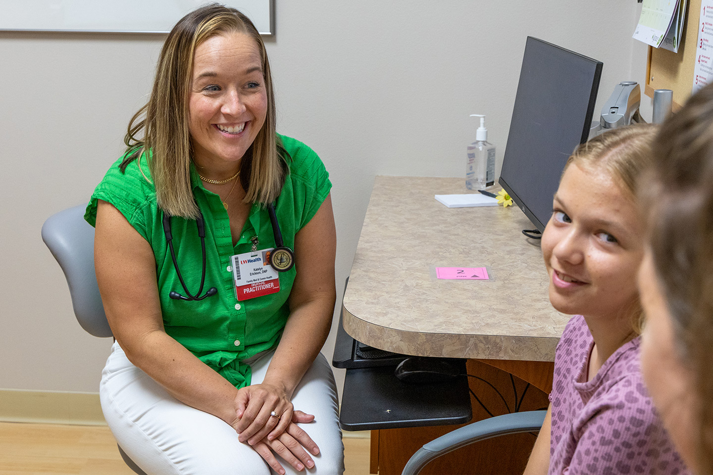 A smiling female doctor in a clinic setting listens to a young girl patient.