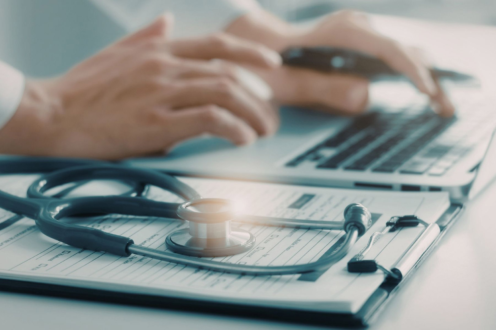 Close up of doctors hands on a keyboard with a stethoscope in the foreground.