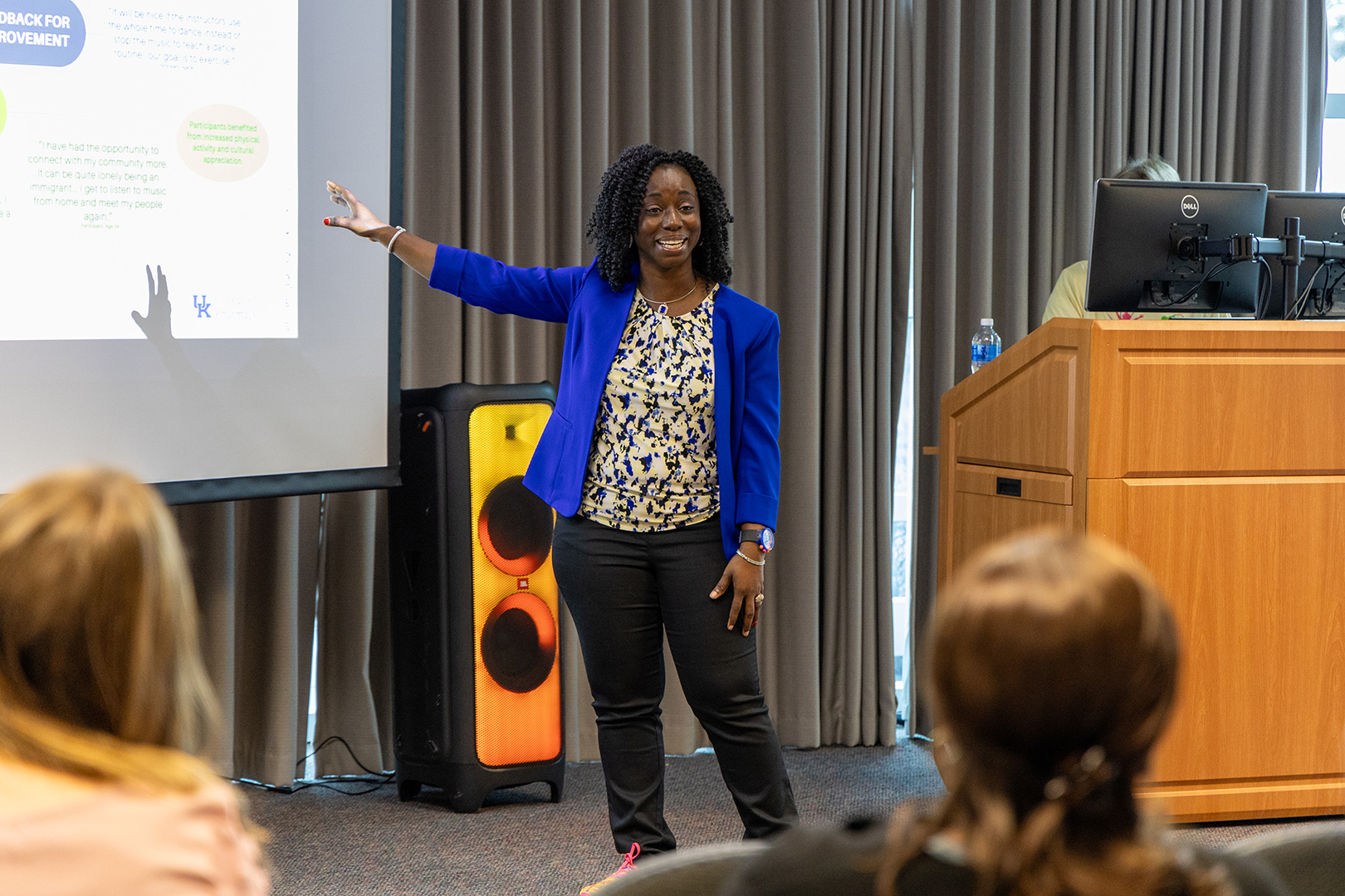 Lecturer standing and gesturing to a screen while presenting to an audience.