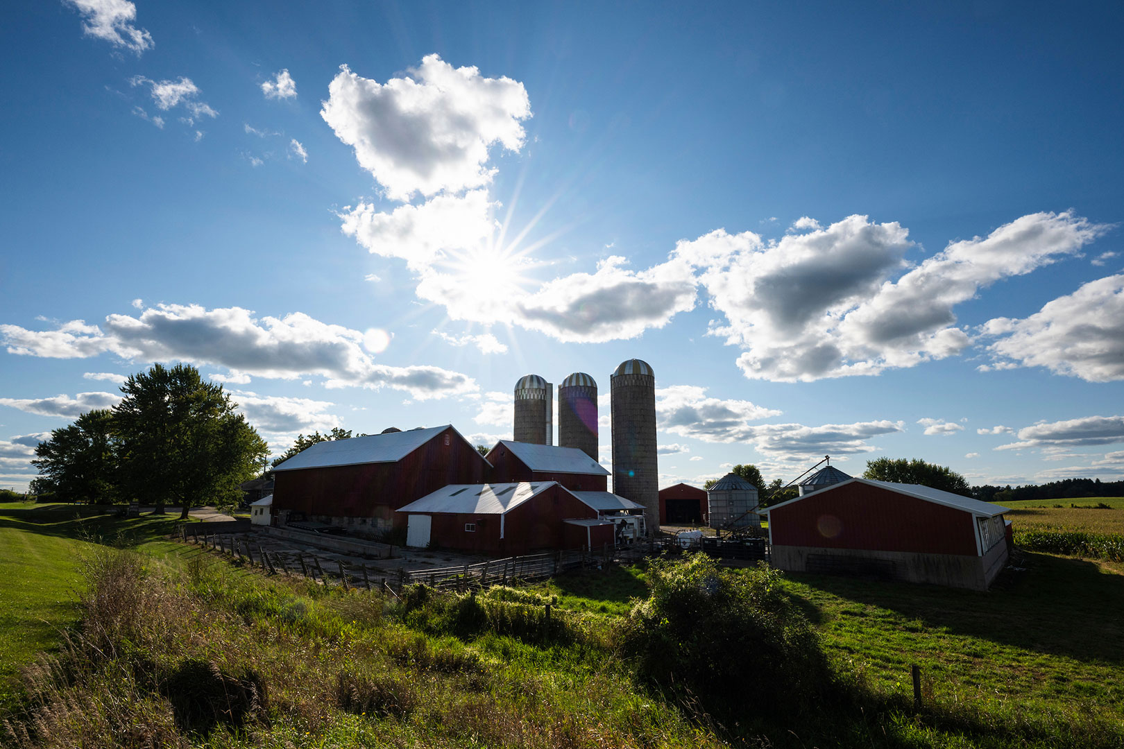 Sunlight burst through puffy clouds in the sky to silhouette a view of rural farmland, including grain silos and a red barn, outside of Beaver Dam, Wis., during a late summer day.