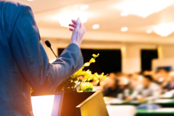 Person standing at podium looking out at room, tightly cropped to show gesturing arm.